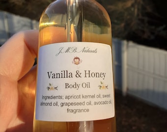 May include: A clear glass bottle with a black dropper top filled with a light amber liquid. The bottle has a white label with the text "J.M.B. Naturals" and "Vanilla & Honey Body Oil". The label also lists the ingredients: apricot kernel oil, sweet almond oil, grapeseed oil, avocado oil, and fragrance.