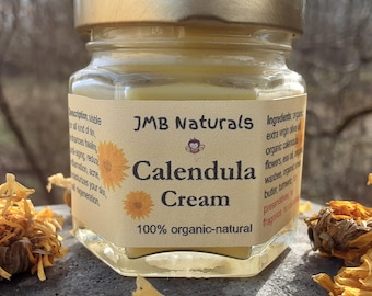 May include: A small glass jar of JMB Naturals Calendula Cream with a gold lid, labeled "100% organic-natural." Dried calendula flowers are scattered around the jar. The cream is a light yellow color.