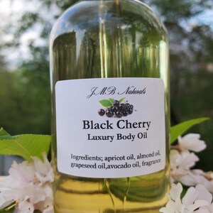 Op de afbeelding: Een heldere glazen fles Black Cherry Luxury Body Oil met een zwarte dop. Het etiket bevat de merknaam, productnaam en ingredi&euml;nten. Witte bloemen en groene bladeren omringen de fles.
