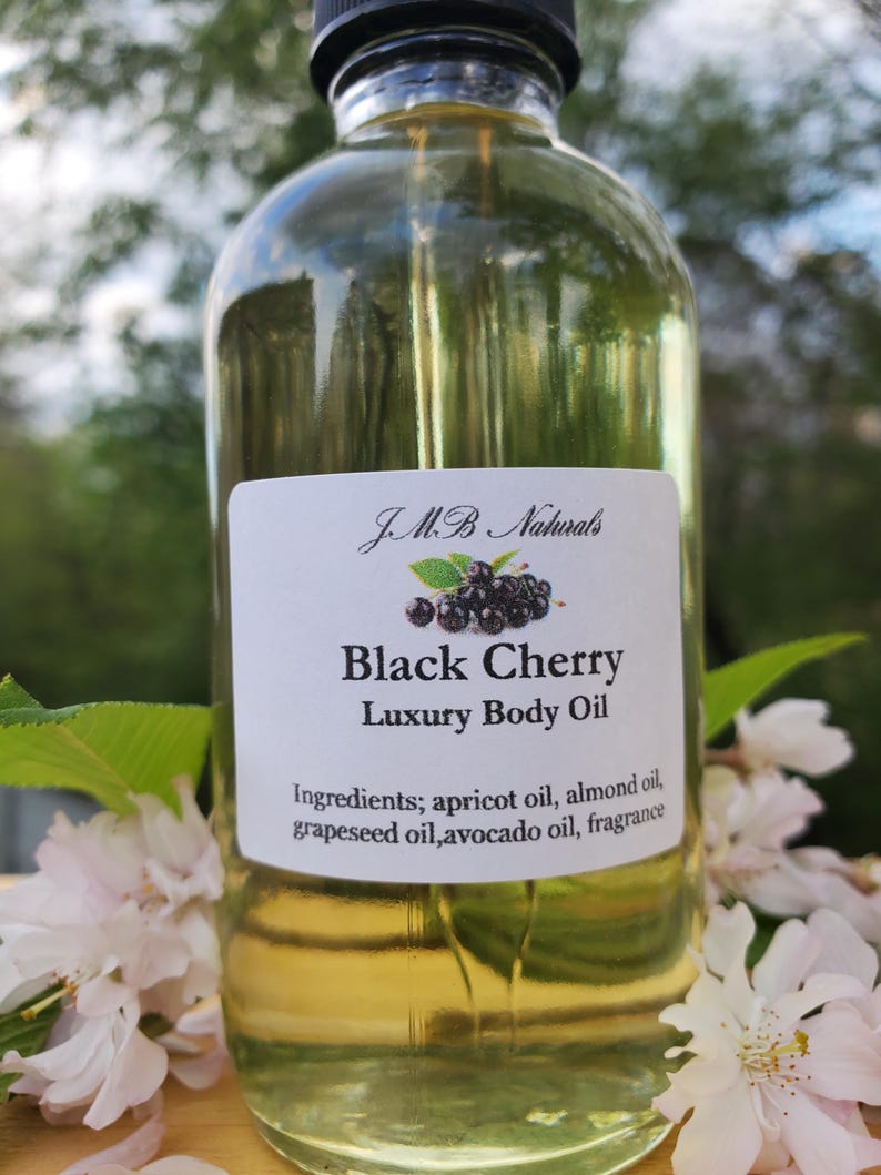 Op de afbeelding: Een heldere glazen fles Black Cherry Luxury Body Oil met een zwarte dop. Het etiket bevat de merknaam, productnaam en ingredi&euml;nten. Witte bloemen en groene bladeren omringen de fles.