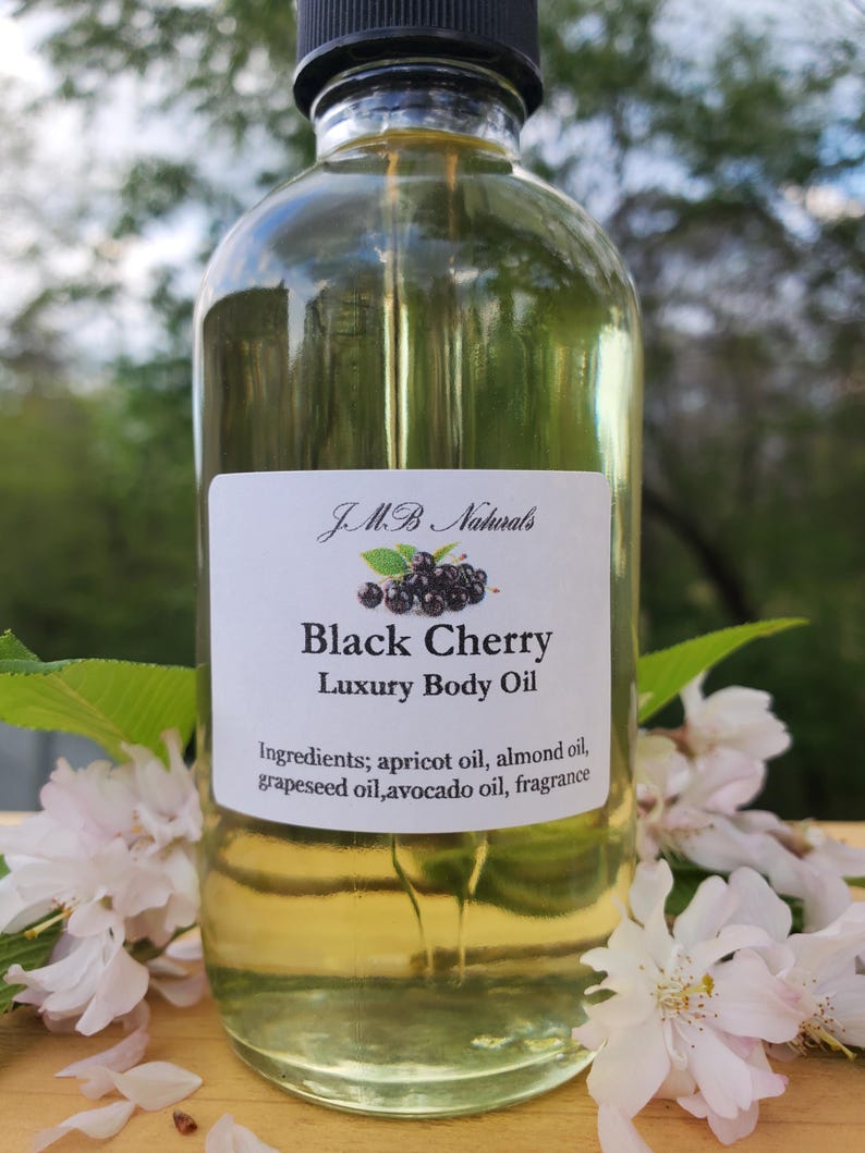 Op de afbeelding: Een heldere glazen fles Black Cherry Luxury Body Oil met een zwarte dop. Het etiket bevat de merknaam, productnaam en ingredi&euml;nten. Witte bloemen en groene bladeren omringen de fles.
