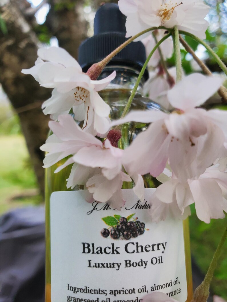 Op de afbeelding: Een heldere glazen fles Black Cherry Luxury Body Oil met een zwarte druppelaar. Het etiket toont een illustratie van zwarte kersen en tekst. Witte en roze kersenbloesems omlijsten de fles.