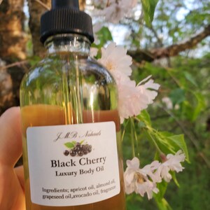 Op de afbeelding: Een heldere glazen fles Black Cherry Luxury Body Oil met een zwarte druppelaar. Het etiket toont de merknaam, productnaam en ingredi&euml;nten. De achtergrond toont witte bloemen en groene bladeren.