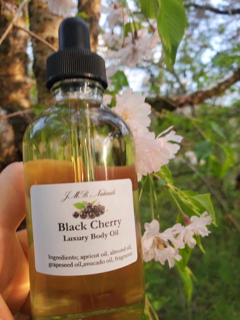 Op de afbeelding: Een heldere glazen fles Black Cherry Luxury Body Oil met een zwarte druppelaar. Het etiket toont de merknaam, productnaam en ingredi&euml;nten. De achtergrond toont witte bloemen en groene bladeren.