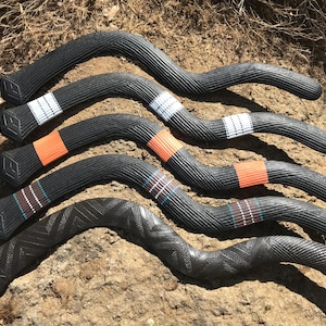 Puede incluir: Varios bumeranes negros con superficies texturizadas. Algunos tienen bandas blancas, naranjas y turquesas. Los bumeranes están colocados sobre un lecho de hojas secas y tierra.