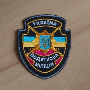Puede incluir: Parche bordado con el escudo de armas ucraniano y texto en ucraniano. El parche tiene un fondo negro con detalles dorados, azules y rosas. El texto incluye "UKRAINA" y "PODATKOVA MILITSIA".