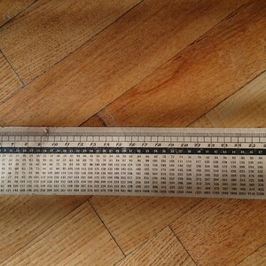 Puede incluir: Una regla rectangular vintage de papel blanquecino, sobre un suelo de madera. La regla está marcada en centímetros hasta 76 cm e incluye una tabla de multiplicar. La regla está ligeramente desgastada.