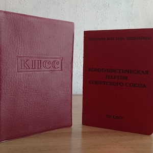 May include: Two burgundy-colored books are displayed on a wooden surface. The book on the left has the embossed Cyrillic letters "КПСС" within a rectangular frame. The book on the right has Cyrillic text.
