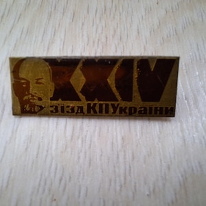 Op de afbeelding: Een rechthoekige speld met een portret van een man aan de linkerkant en de letters "XXIV" aan de rechterkant. De speld heeft tekst in een vreemde taal aan de onderkant. De speld is goud en bruin.