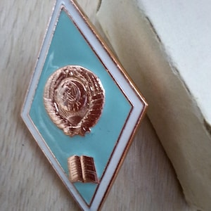 Pode incluir: Um alfinete em forma de losango com um centro azul claro, borda branca e detalhes em cobre. O alfinete apresenta um brasão de armas e um livro aberto. O alfinete está ao lado de uma caixa de papelão.