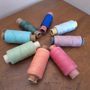 May include: A collection of eight vintage sewing thread spools arranged in a circular pattern. The spools feature various colors including dark green, bright blue, light blue, pale pink, dusty rose, coral, light green, and turquoise on cardboard cores.