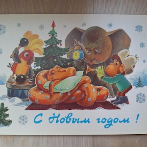 Puede incluir: Postal navideña vintage con animales de dibujos animados celebrando. Una serpiente naranja descansa sobre una almohada, mientras un elefante, un mono y un loro tocan instrumentos. El texto "Feliz Año Nuevo" está escrito en azul.