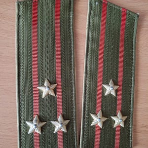 May include: A pair of olive green military shoulder boards featuring three vertical red stripes and three gold-colored metal stars on each piece. The rectangular fabric straps have a textured weave and are designed for uniform attachment.
