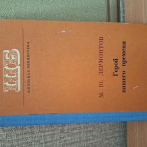 Può includere: Un libro d'epoca intitolato "Eroe del nostro tempo" di M. Yu. Lermontov. Il libro ha una copertina arancione con bordi blu e testo in russo. La copertina presenta anche il logo della "Biblioteca scolastica".