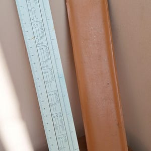 May include: A vintage slide rule with a white and black scale, and a brown leather case. The slide rule has various markings and numbers, and the case has text on it. A classic tool for calculations.