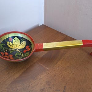 May include: A wooden ladle featuring a round bowl with a black interior painted with a gold leaf design and red berries. The handle is painted in sections of red and gold, with a curved end. The exterior of the bowl is painted in red.