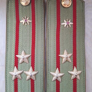 May include: Pair of military shoulder boards in olive green with red stripes. Each board features gold-colored star-shaped insignia and a circular button at the top. The boards are likely for a military uniform.