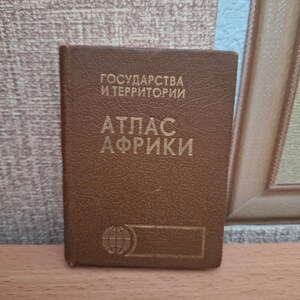 May include: A brown atlas with gold lettering. The title reads "ATLAS AFRIKI" in Cyrillic script, with additional text above. A globe icon is visible near the bottom. The book is rectangular and appears to be made of leather or a similar material.