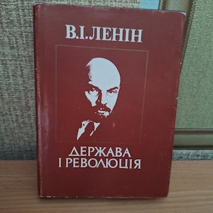 May include: A maroon book with the title "V.I. Lenin" in white lettering. Below the title is a white portrait of a man with a beard. The text "ДЕРЖАВА I РЕВОЛЮЦІЯ" is printed in white at the bottom of the book.