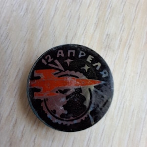 May include: A round, black pin with the text "12 АПРЕЛЯ" in white and a red and orange rocket design. The pin features a globe and stars, likely commemorating a historical event.