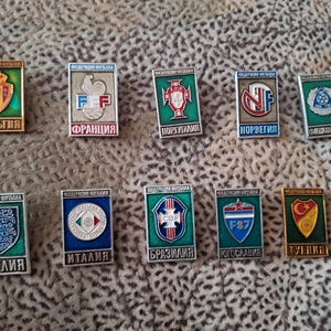 Peut inclure: Une collection de 10 épingles en émail représentant les armoiries des équipes nationales de football de Belgique, France, Portugal, Norvège, Finlande, Angleterre, Italie, Brésil, Yougoslavie et Turquie. Chaque épingle est verte, blanche et rouge avec une bordure dorée et le nom du pays en cyrillique.