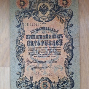 Puede incluir: Un billete antiguo de cinco rublos de 1909, con diseños florales y de volutas ornamentados. El billete tiene el texto "ГОСУДАРСТВЕННЫЙ КРЕДИТНЫЙ БИЛЕТЬ ПЯТЬ РУБАВИ" y el número "5" en varios lugares.