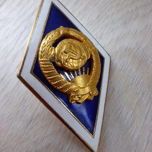 Könnte beinhalten: Eine rautenförmige Anstecknadel mit einem goldenen Emblem auf blauem Hintergrund, umrahmt von einem weißen Rand. Das Emblem zeigt eine Weltkugel, Hammer und Sichel, umgeben von einem Kranz. Die Nadel hat einen goldfarbenen Rand.