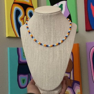 May include: A white beaded necklace with blue, orange, and white beads. The necklace is on a white mannequin bust.