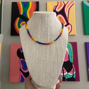 May include: A rainbow beaded necklace on a white mannequin bust. The necklace is made of small, colorful beads in the order of red, orange, yellow, green, blue, indigo, and violet.