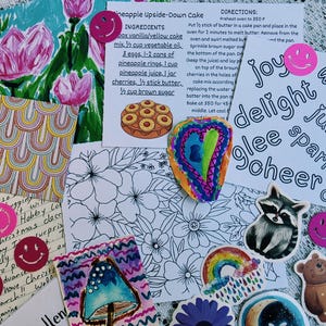 May include: A collection of paper ephemera including a pineapple upside-down cake recipe card, a floral coloring page, a handwritten letter, and various stickers featuring a raccoon, a bear, rainbows, mushrooms, and pink smiley faces.