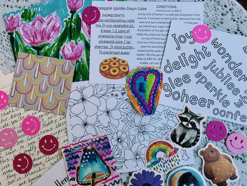 May include: A collection of paper ephemera including a pineapple upside-down cake recipe card, a floral coloring page, a handwritten letter, and various stickers featuring a raccoon, a bear, rainbows, mushrooms, and pink smiley faces.