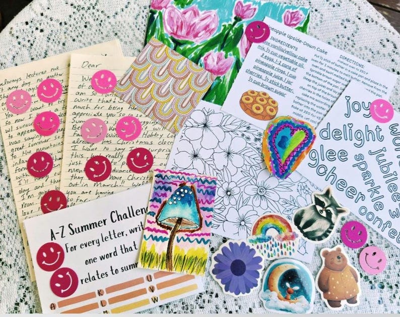May include: A collection of stationery items including handwritten letters, a recipe card for pineapple upside-down cake, a floral coloring page, a mushroom illustration, and various stickers featuring smiley faces, a raccoon, a bear, and a rainbow.
