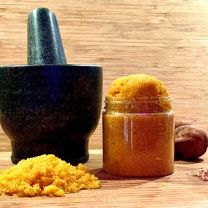 Puede incluir: Un tarro de vidrio lleno de un exfoliante de azúcar amarillo-naranja, un mortero y un pilón, y una nuez marrón sobre una superficie de madera.
