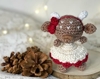 May include: A crocheted reindeer toy wearing a white dress with red trim. The reindeer has brown yarn for its body and red yarn for its nose and antlers.