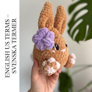 Puede incluir: Un conejito de ganchillo marrón claro con una flor lavanda. El conejito tiene patas rosas y blancas y ojos negros. El texto en el lado izquierdo de la imagen dice "ENGLISH US TERMS - SVENSKA TERMER".