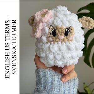 May include: A handmade, white, plush sheep toy with a light pink flower and tan ears. The sheep has a tan face with black eyes and a pink nose. The text on the left side of the image reads "ENGLISH US TERMS - SVENSKA TERMER".