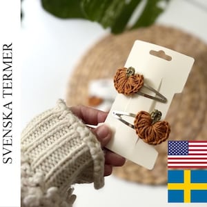 May include: Two silver-toned metal snap hair clips attached to a white display card. Each clip features a small, hand-crocheted pumpkin ornament in a warm orange yarn with a tiny green stem detail, showcasing a textured, circular design.