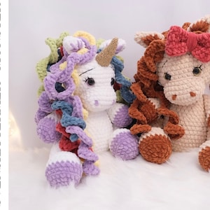 May include: Two crocheted unicorn plushies, one white with a rainbow mane and a brown horn, and one brown with a pink bow. The plushies are sitting on a white fluffy surface.