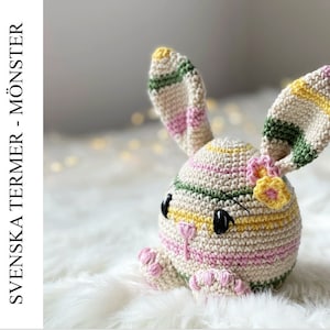 May include: A crocheted bunny toy with pink, yellow, and green stripes. The bunny has black eyes and a small flower on its head.