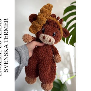 以下が含まれることがあります： 手作りの、茶色の牛のぬいぐるみ。薄茶色の鼻面、ひづめ、アクセントがあります。牛の頭には、大きくて質感のある薄茶色のリボンが付いています。「ENGLISH US TERMS - SVENSKA TERMER」というテキストが左側にあります。