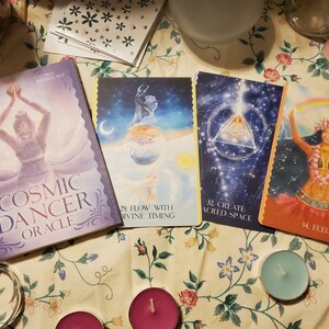 以下が含まれることがあります： Cosmic Dancer Oracleのカードデッキの箱と、天体の設定で人物が描かれた3枚のカード。カードには「29 Flow with Divine Timing」、「32 Create Sacred Space」、「34 Feel」と記されており、箱のタイトルもはっきりと確認できます。