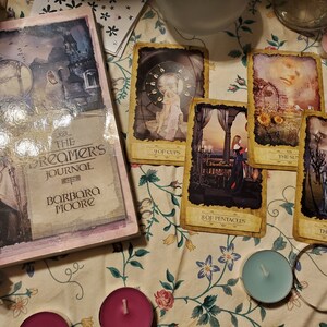 May include: A book titled The Dreamer's Journal by Barbara Moore alongside four tarot cards labeled 9 of Cups, The Sun, 8 of Pentacles, and The Lovers. The book cover features a fantasy illustration with a castle, and the cards display detailed artwork.