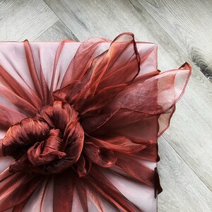Pode incluir: Uma caixa de presente quadrada embrulhada em papel rosa pálido e adornada com um laço grande e elaborado feito de tecido vermelho escuro transparente. O laço é centrado na caixa e tem uma aparência texturizada e em camadas.
