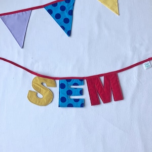 Könnte beinhalten: Ein farbenfrohes Banner mit den Buchstaben "SEM" in Gelb, Blau und Rot. Der blaue Buchstabe hat ein Tupfenmuster. Das Banner ist an einem roten Band befestigt, mit zusätzlichen Wimpelfahnen in Lila, Blau und Gelb.