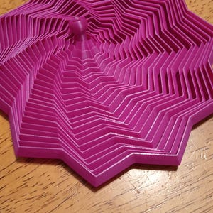 May include: A magenta-colored, star-shaped trivet with a layered, geometric design. The trivet has a small, rounded handle in the center and is made of a rigid material. It sits on a wooden surface.