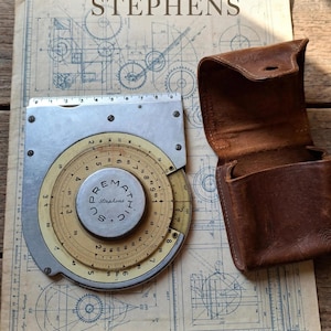 May include: A vintage Supremathic Stephens circular slide rule with a metallic body and a cream-colored rotating dial featuring numerical scales. The device is accompanied by a brown leather carrying case and rests on a technical blueprint.