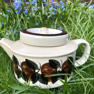 Peut inclure: Une théière en céramique blanche avec un couvercle et une poignée marrons. La théière est décorée d'un motif floral brun et vert avec des feuilles et des fruits. La théière est posée sur un lit d'herbe verte.
