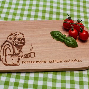 Könnte beinhalten: Ein Holzbrett mit einem Skelett, das eine Tasse Kaffee hält, und dem Text "Kaffee macht schlank und schön" auf der Oberfläche. Auf dem Brett liegen rote Tomaten und grüne Basilikumblätter.