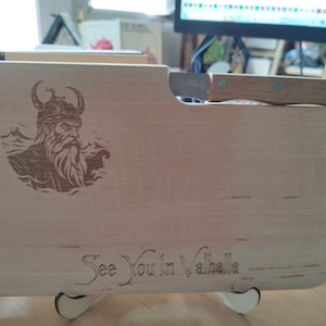 Könnte beinhalten: Ein Holzbrett zum Schneiden mit einem Wikingerkrieger-Design und dem Text "See You In Valhalla". Das Brett hat einen integrierten Messerhalter und ein kleines Messer befindet sich im Halter.