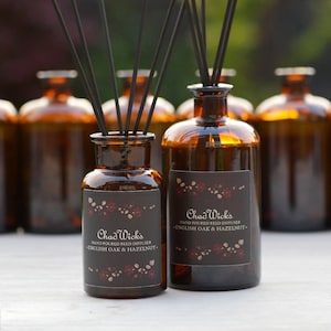 May include: Two amber glass bottles with black reed diffusers and labels that read "Chad Wicks Hand Poured Reed Diffuser - English Oak & Hazelnut - ".